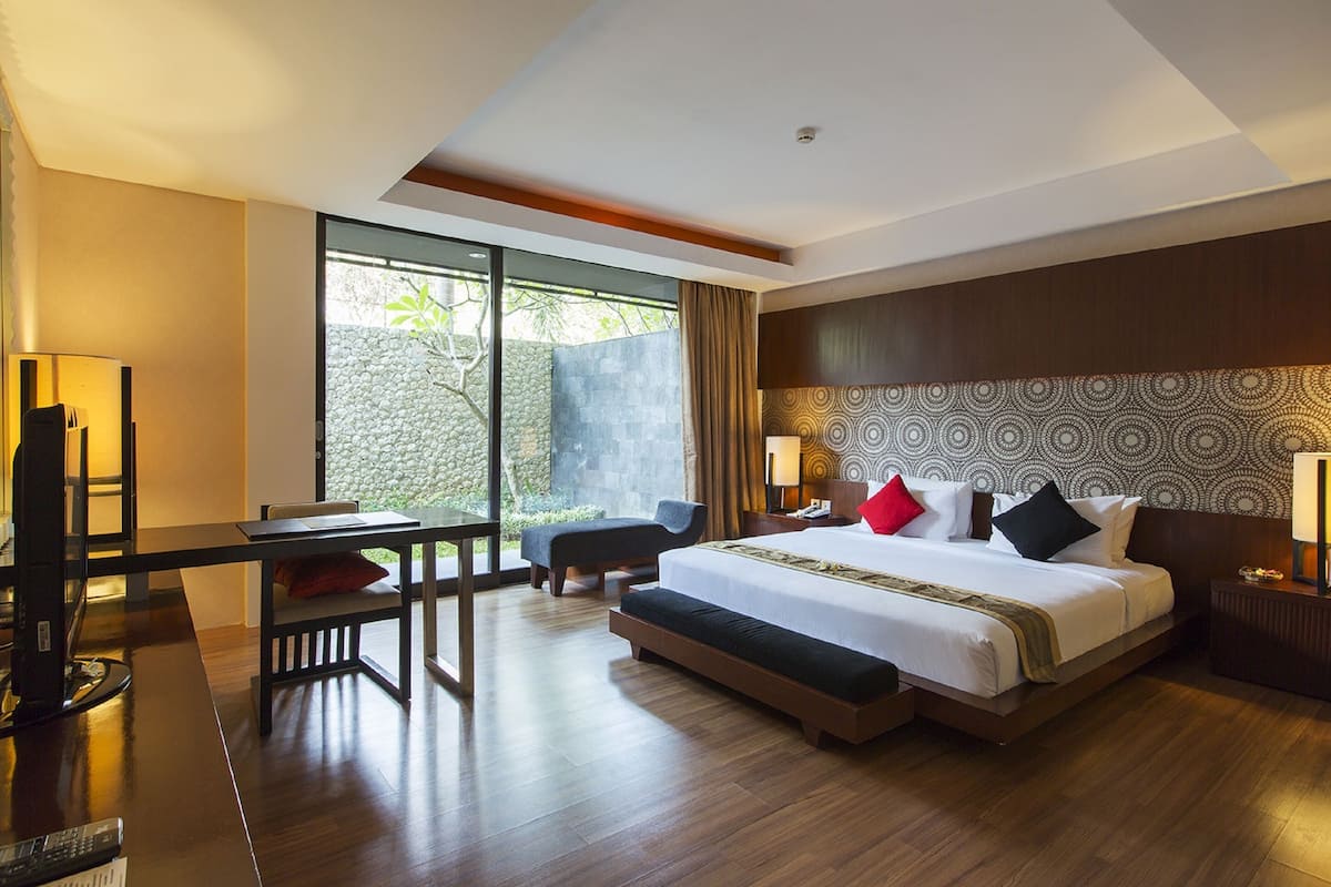 A hotel room with a large bed, a desk with a chair, a TV, and a window with a view of greenery.