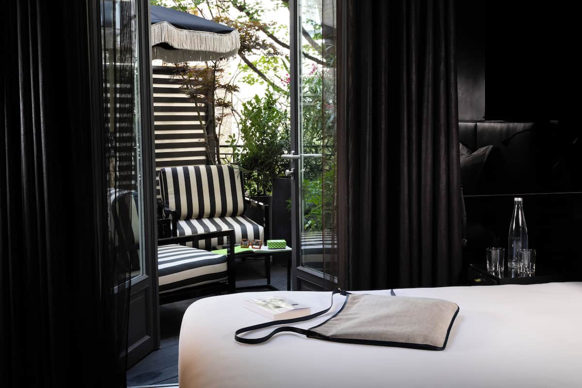 Une chambre d’hôtel avec un lit, un fauteuil à rayures, une petite table avec un plateau vert et une vue sur l’extérieur grâce à de grandes fenêtres.