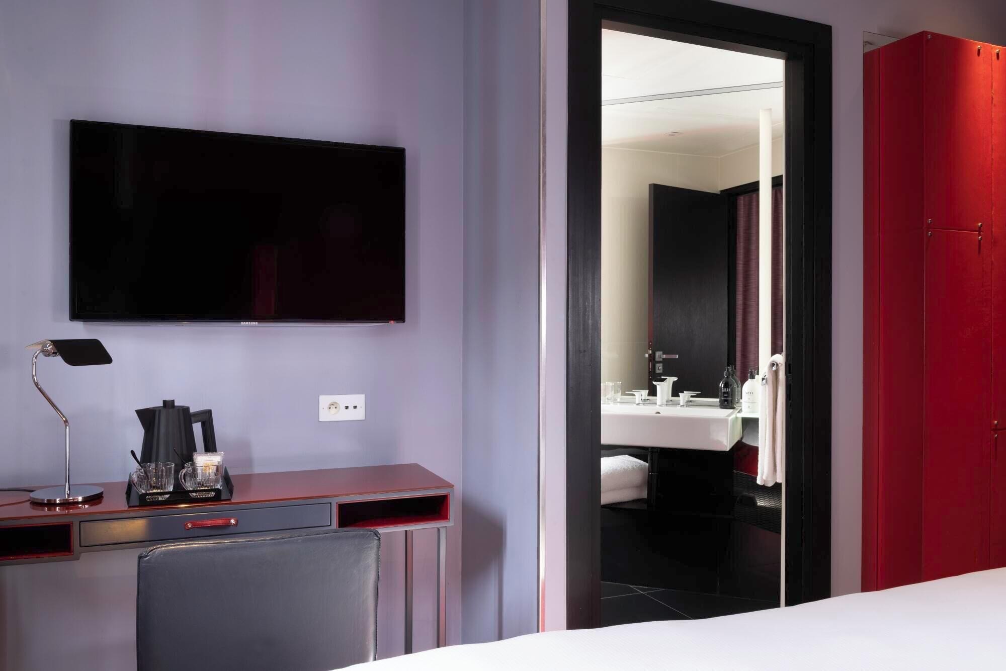 Ein Hotelzimmer mit Schreibtisch, Flachbildfernseher, Spiegel und einem roten Kleiderschrank.