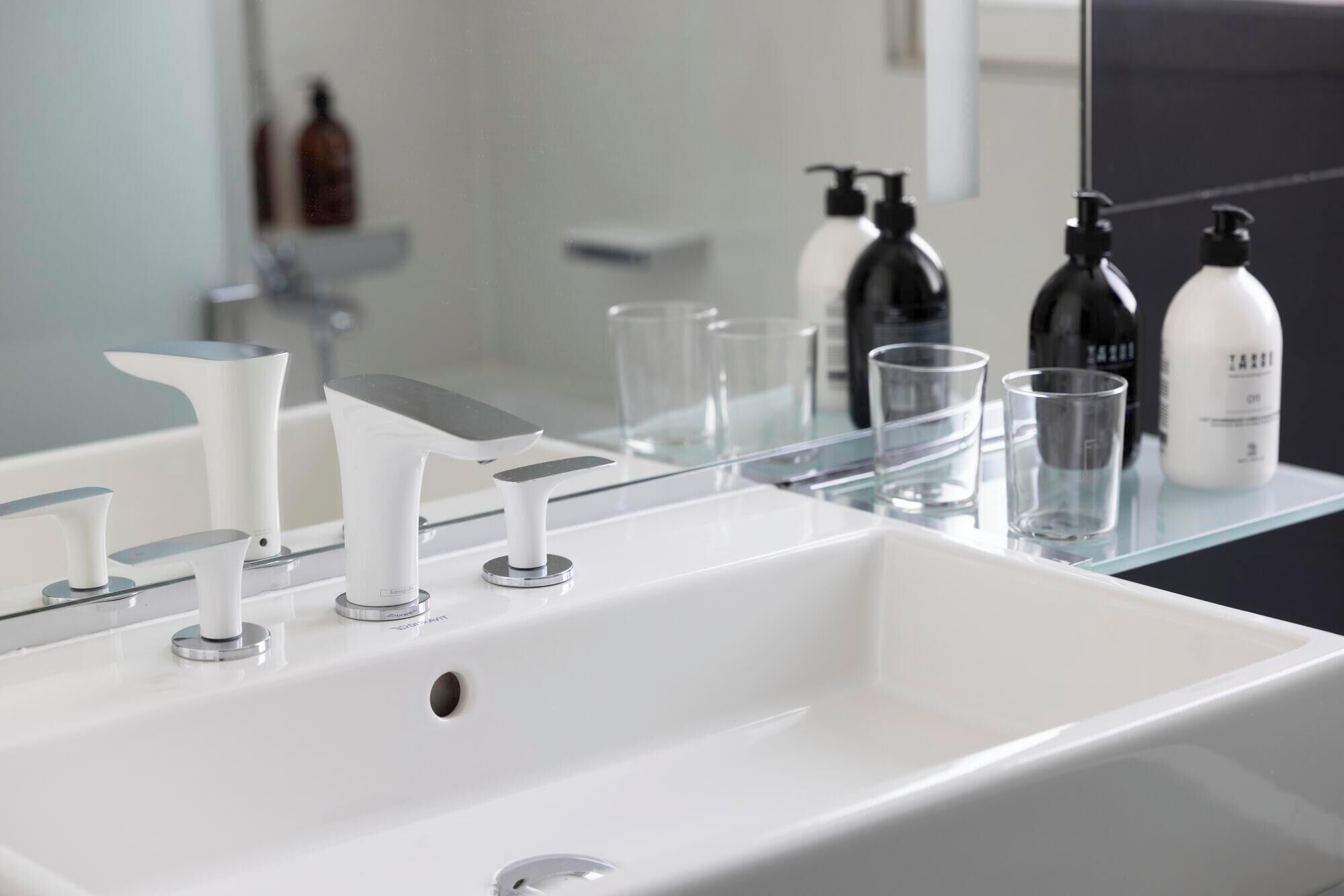 A modern bathroom with a white sink, two faucets, and a glass shelf holding toiletries and glasses.