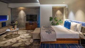 Habitación de hotel moderna con una cama grande, un sofá, un televisor, una mesita de centro y una mesita auxiliar pequeña.
