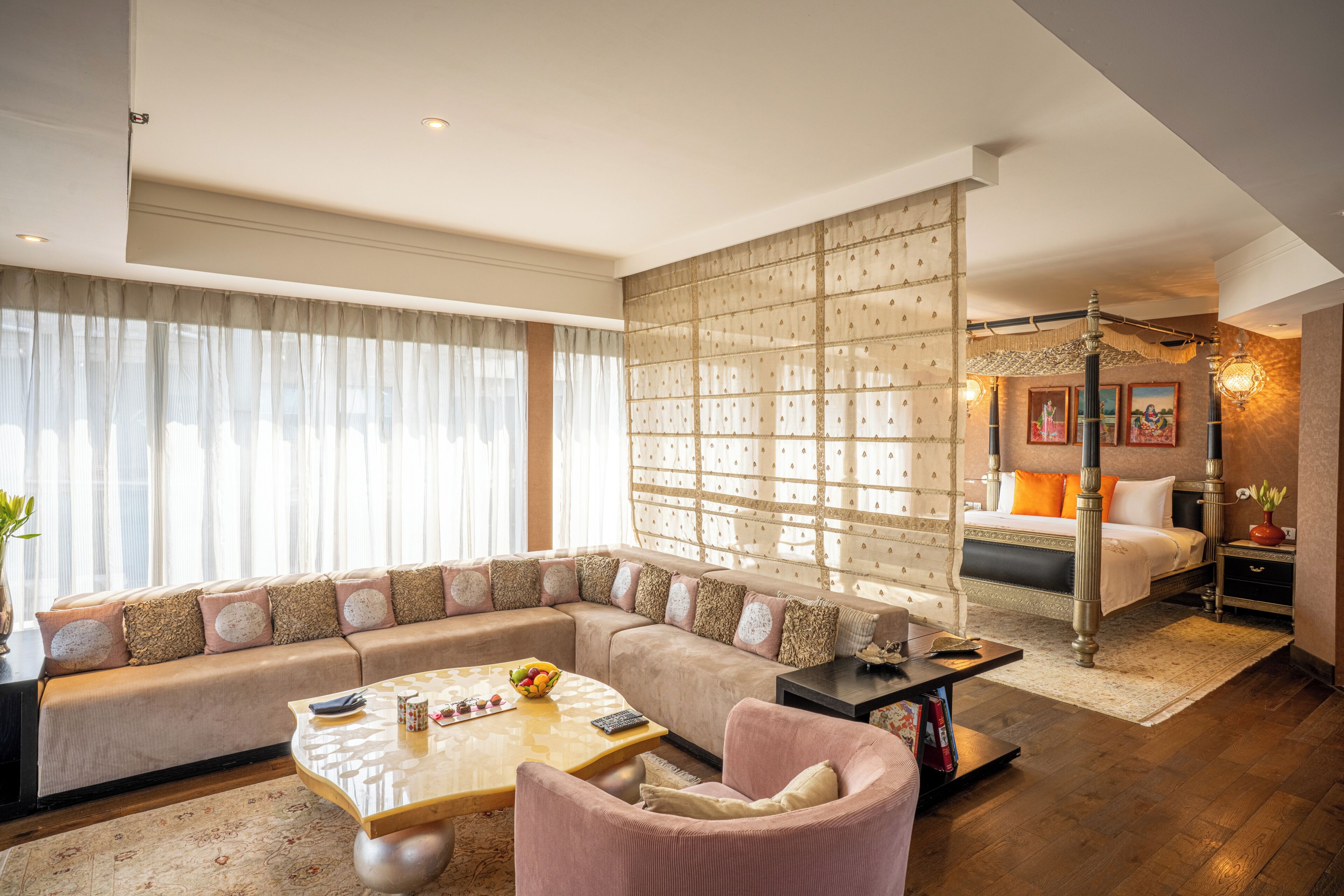 A modern hotel room with a large sofa, a separate seating area, a bed with an ornate headboard, and a decorative wall partition.