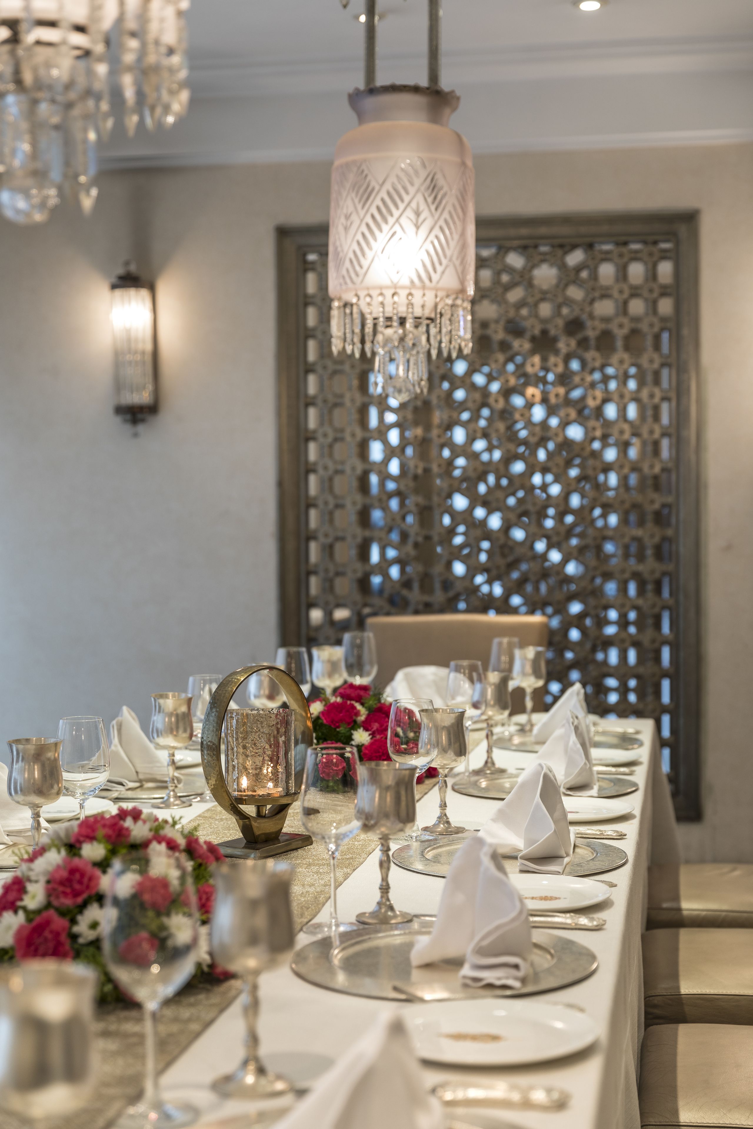 Una larga mesa de comedor vestida con manteles blancos, cubiertos y cristalería, con un centro de mesa decorativo y un lámpara colgante arriba.