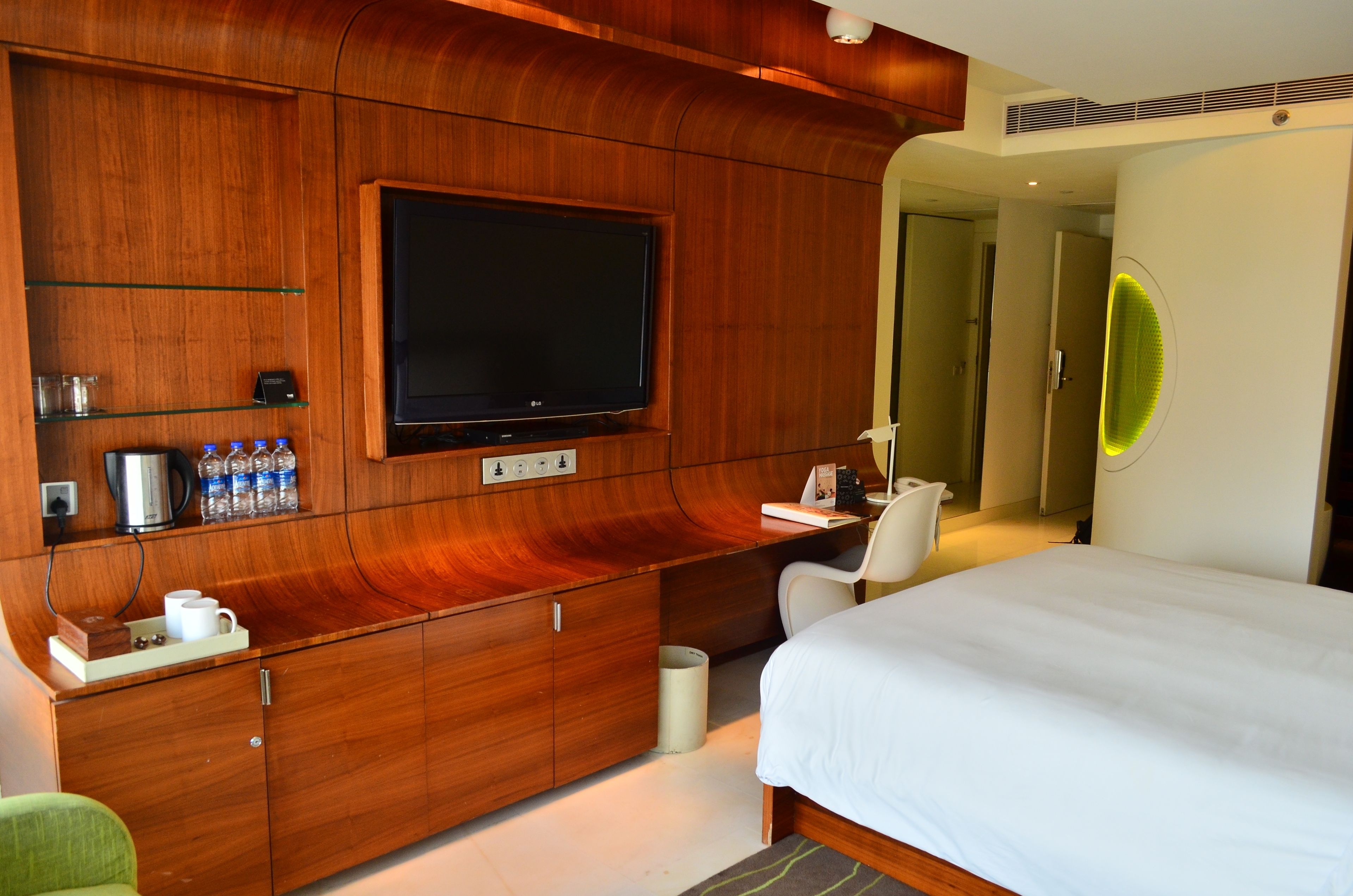 A hotel room with a large flat-screen TV mounted on a wooden wall, a bed with white bedding, a desk with a chair, and a mirror with a neon light.