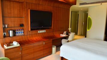 A hotel room with a large flat-screen TV mounted on a wooden wall, a bed with white bedding, a desk with a chair, and a mirror with a neon light.