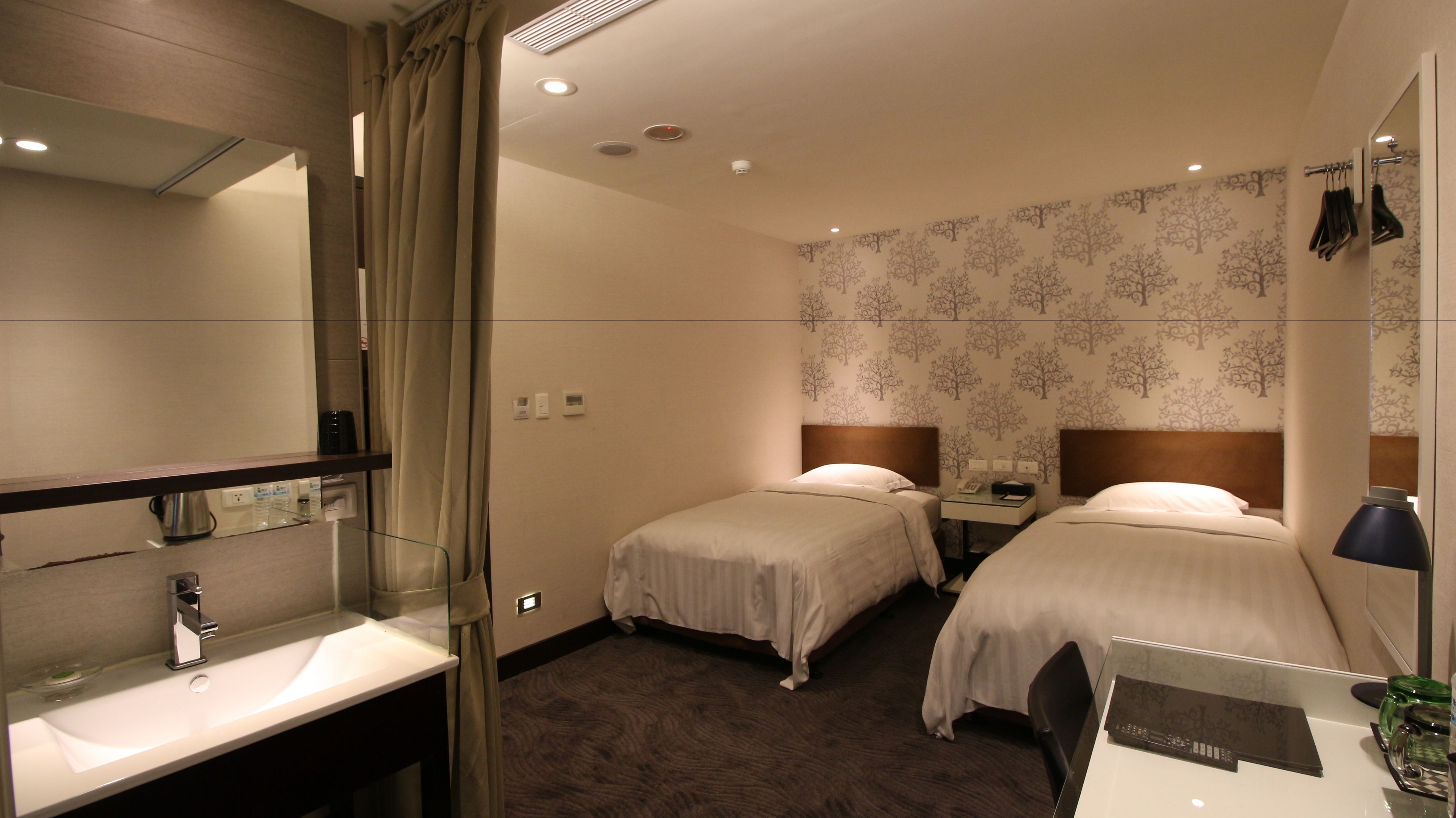A hotel room with two single beds, a bathroom with a sink and mirror, and a small desk with a telephone and a lamp.