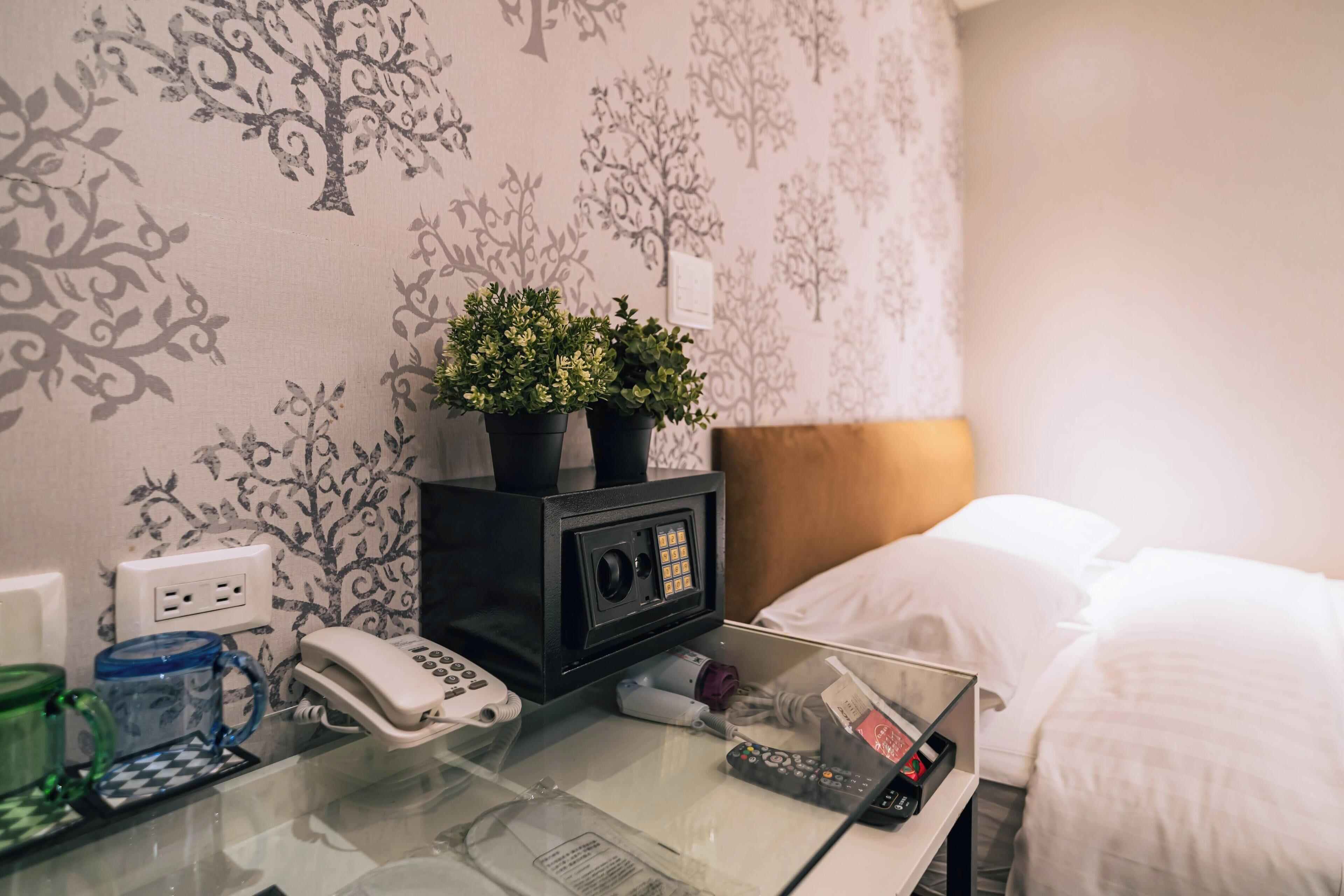 A hotel room with a bed, a bedside table with a phone, a small plant, and a wall with a tree pattern.