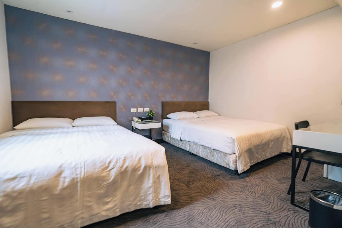 A hotel room with two beds, a desk, a chair, a nightstand with a phone and a plant, and a patterned wall.
