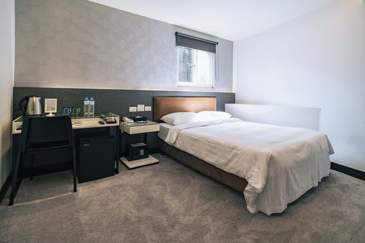 A hotel room with a bed, a desk with a chair, a nightstand, a window with a view, and a wall-mounted radiator.