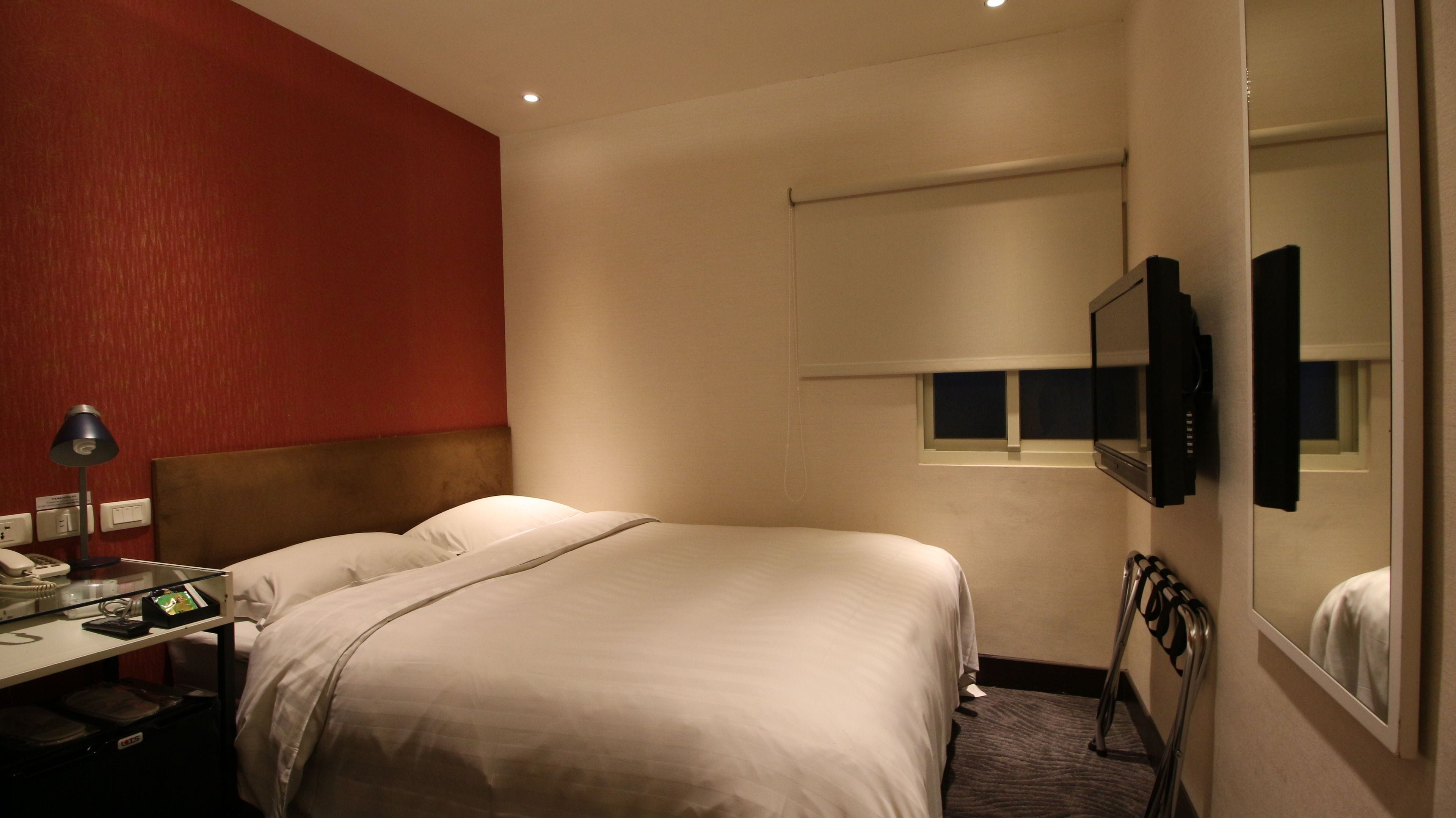 A hotel room with a bed, bedside table, lamp, and a television mounted on the wall.