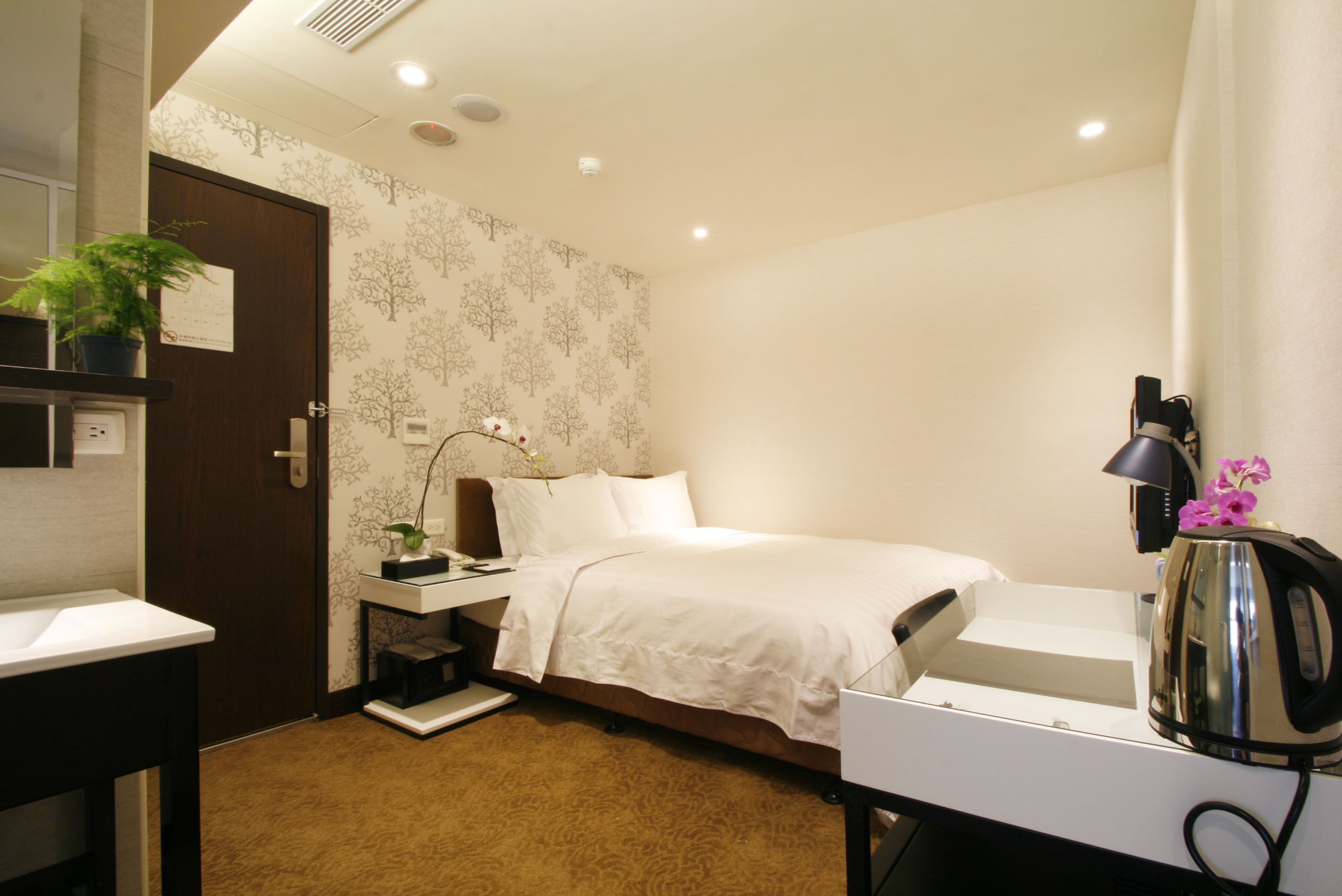 Een hotelkamer met een bed, nachtkastje, lamp en een waterkoker.