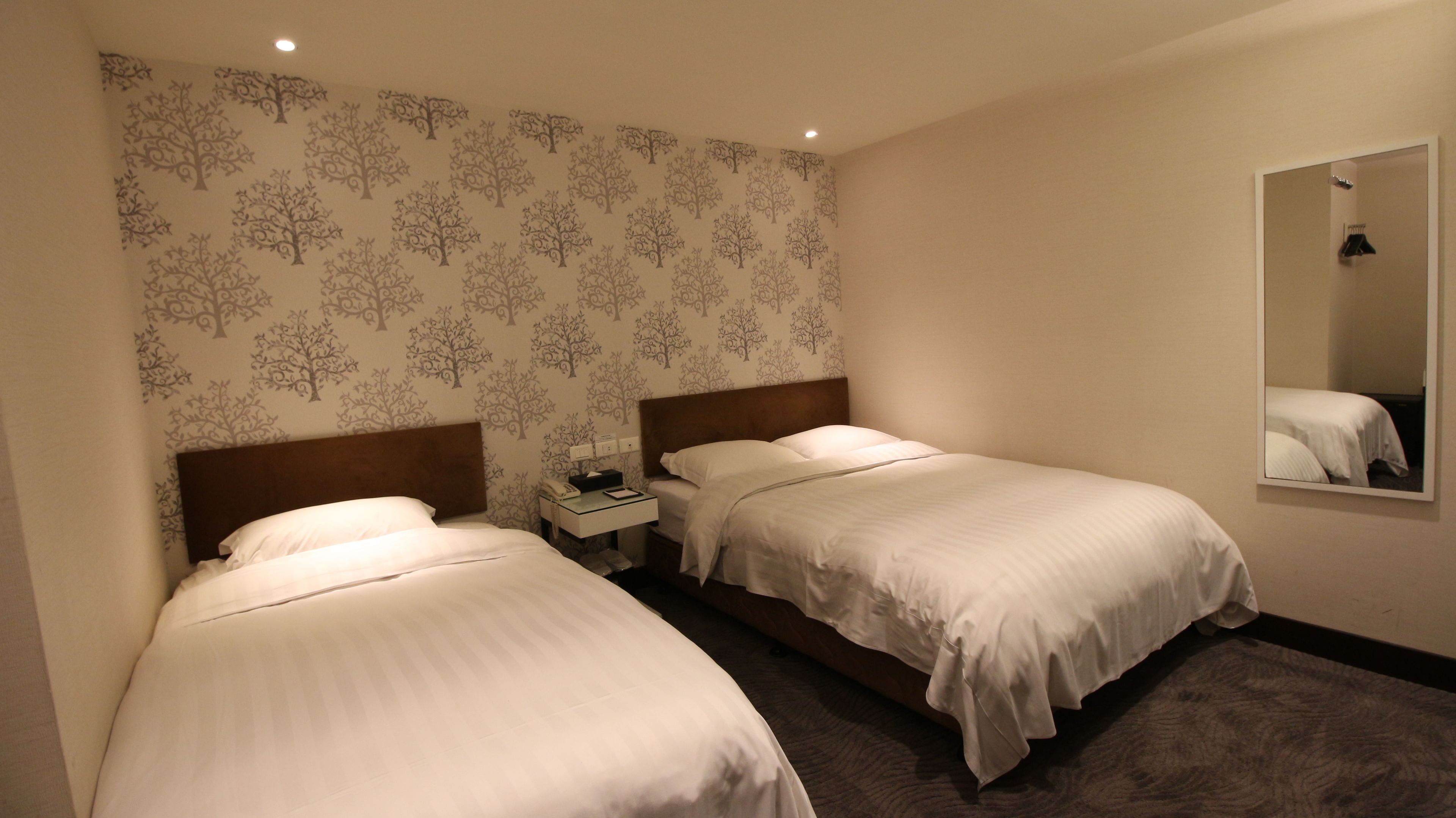 Habitación de hotel con dos camas individuales, una mesita de noche, un espejo y un papel tapiz con un diseño de árbol.