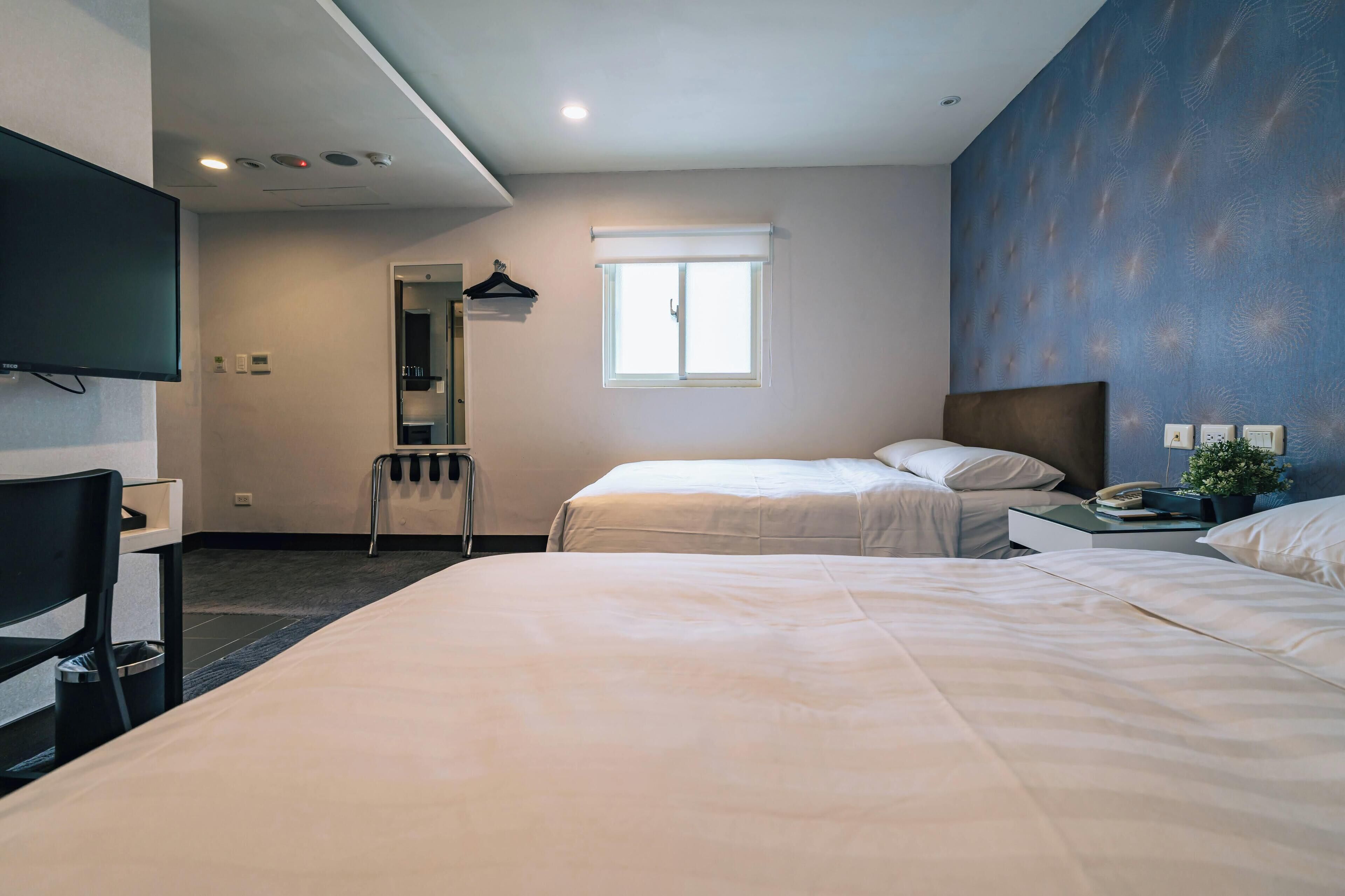 A hotel room with two beds, a desk, a chair, a TV, a window, and a mirror.
