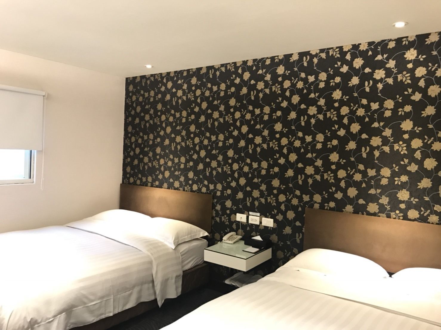 Ein Hotelzimmer mit zwei Betten, einem kleinen Nachttisch mit Telefon und einem Fenster mit Jalousien.
