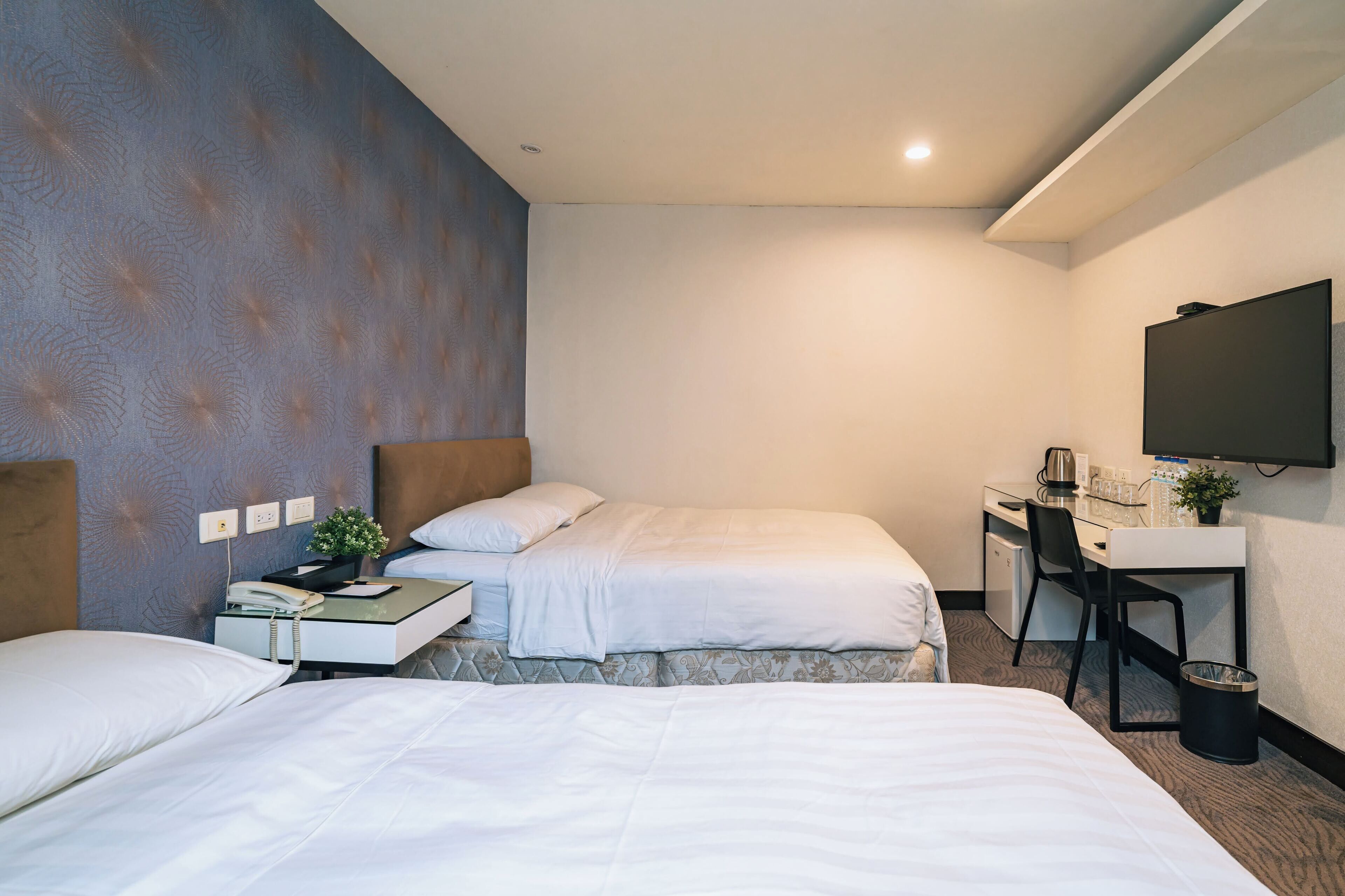 A hotel room with two beds, a desk, a chair, a TV, and a small trash bin.