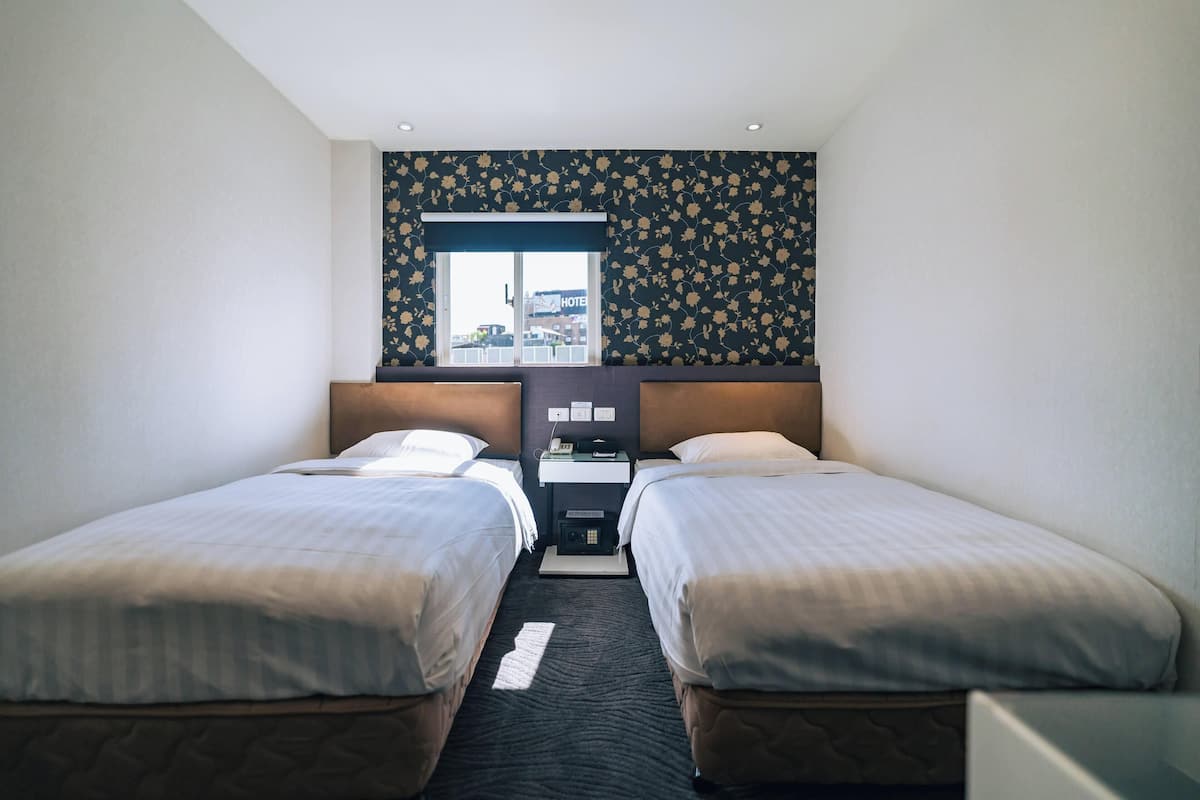 A hotel room with two beds, a small desk with a telephone, and a window with a view of the outside.