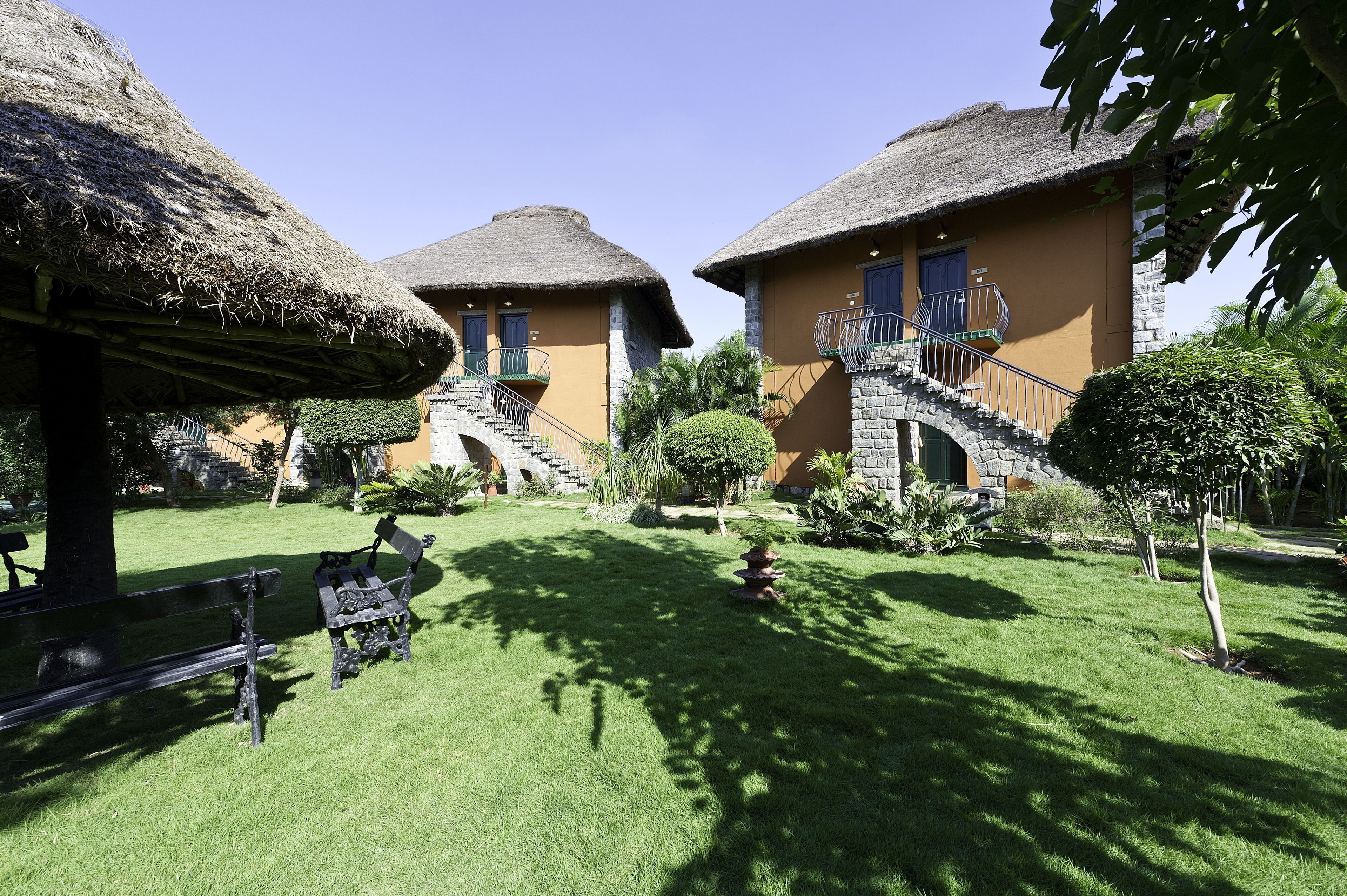 A courtyard with two thatched-roof buildings, a bench, and a small tree.