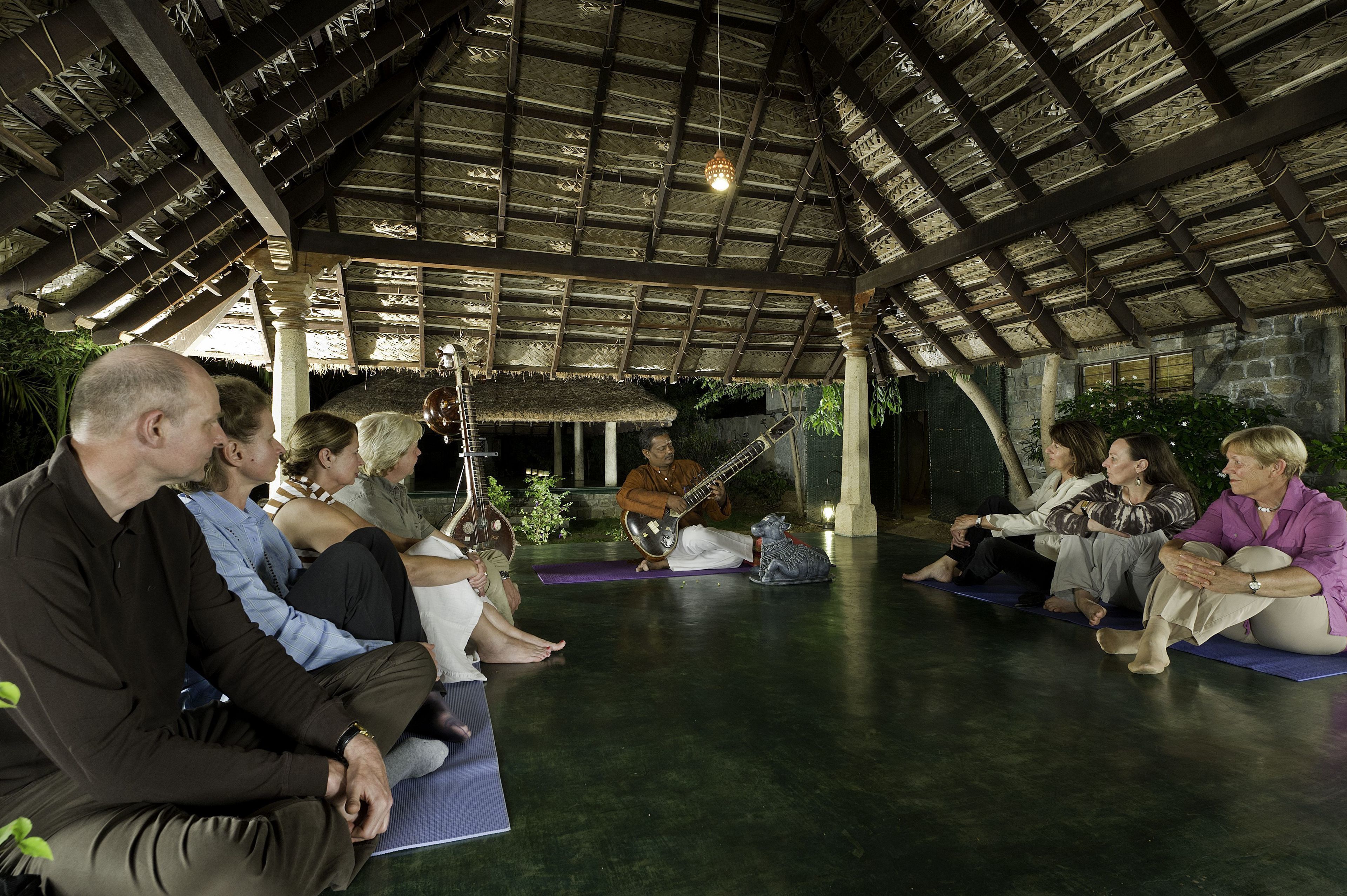 Un grupo de personas sentadas sobre esterillas de yoga en un espacio cubierto con techo de paja y paredes de piedra.