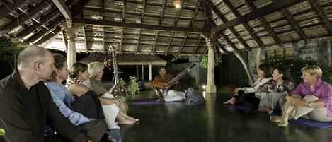 Un grupo de personas sentadas sobre esterillas de yoga en un espacio cubierto con techo de paja y paredes de piedra.