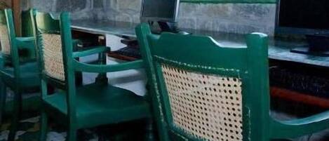 A room with a stone wall, a window with a view of a building, a table with a computer, and green chairs.