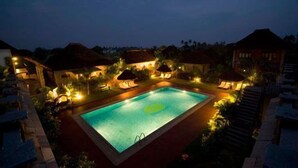 Une piscine rectangulaire entourée de cabanes aux toits de chaume et de chaises longues, avec un espace piscine bien éclairé au crépuscule.