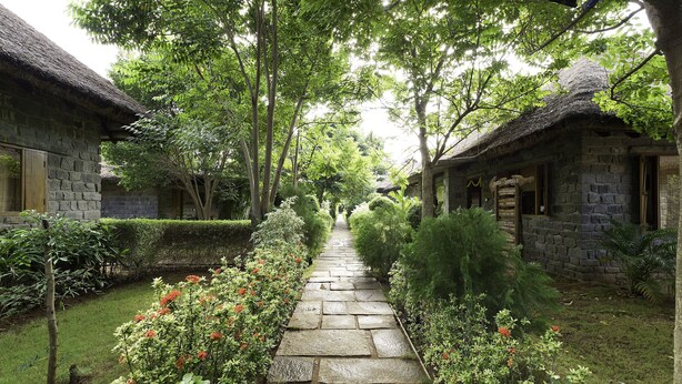 Un sentier de pierre mène à des bâtiments traditionnels en pierre, entourés d’une végétation luxuriante et de plantes en fleurs.