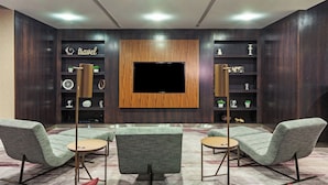 Un salon moderne avec un téléviseur à écran plat fixé sur un panneau en bois, deux fauteuils rembourrés, une petite table ronde et une bibliothèque agrémentée d’objets décoratifs.