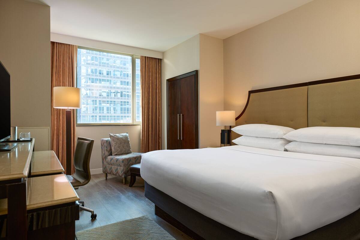 Quarto de hotel com uma cama grande, uma escrivaninha, uma cadeira, um guarda-roupa e uma janela com cortinas.