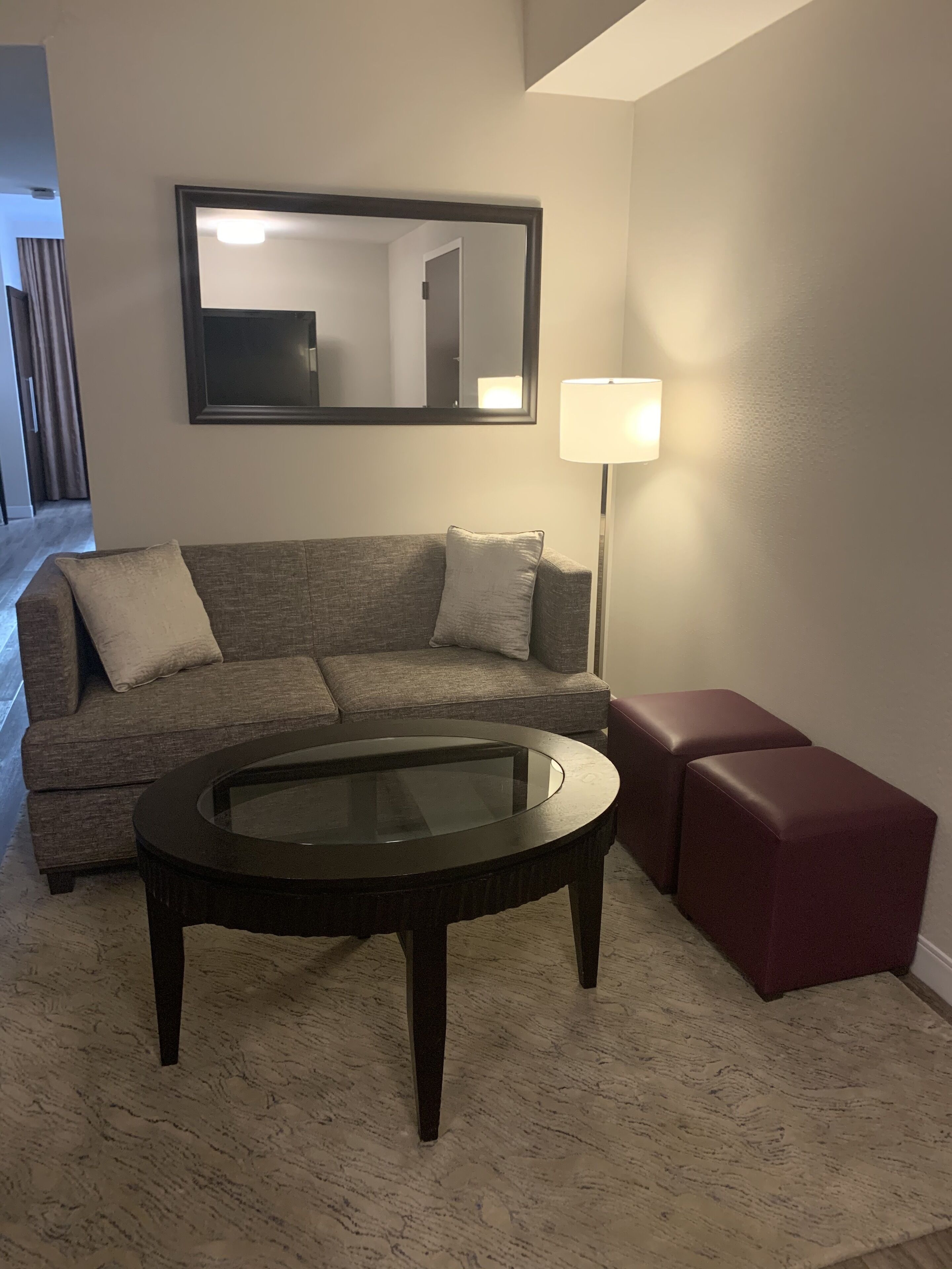 A living room with a sofa, a glass-top coffee table, a floor lamp, and a red ottoman.