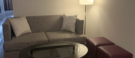 Una sala de estar con un sofá, una mesa de centro de vidrio, una lámpara de pie y un reposapiés rojo.