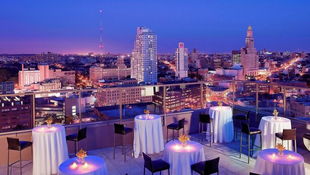 A rooftop terrace with round tables and chairs, set against a cityscape at dusk.
