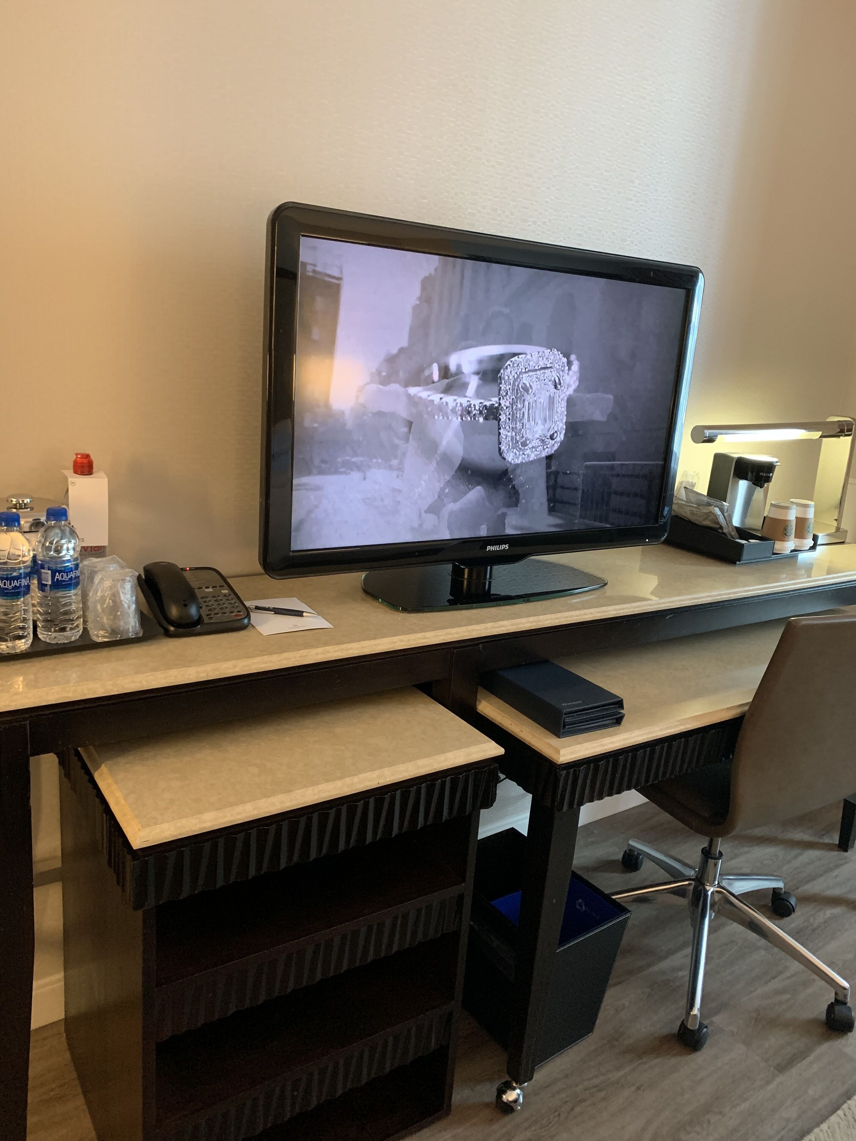 A hotel room with a desk featuring a computer monitor, a phone, and a keyboard. There are water bottles, a coffee maker, and a trash bin on the desk.