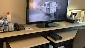 A hotel room with a desk featuring a computer monitor, a phone, and a keyboard. There are water bottles, a coffee maker, and a trash bin on the desk.