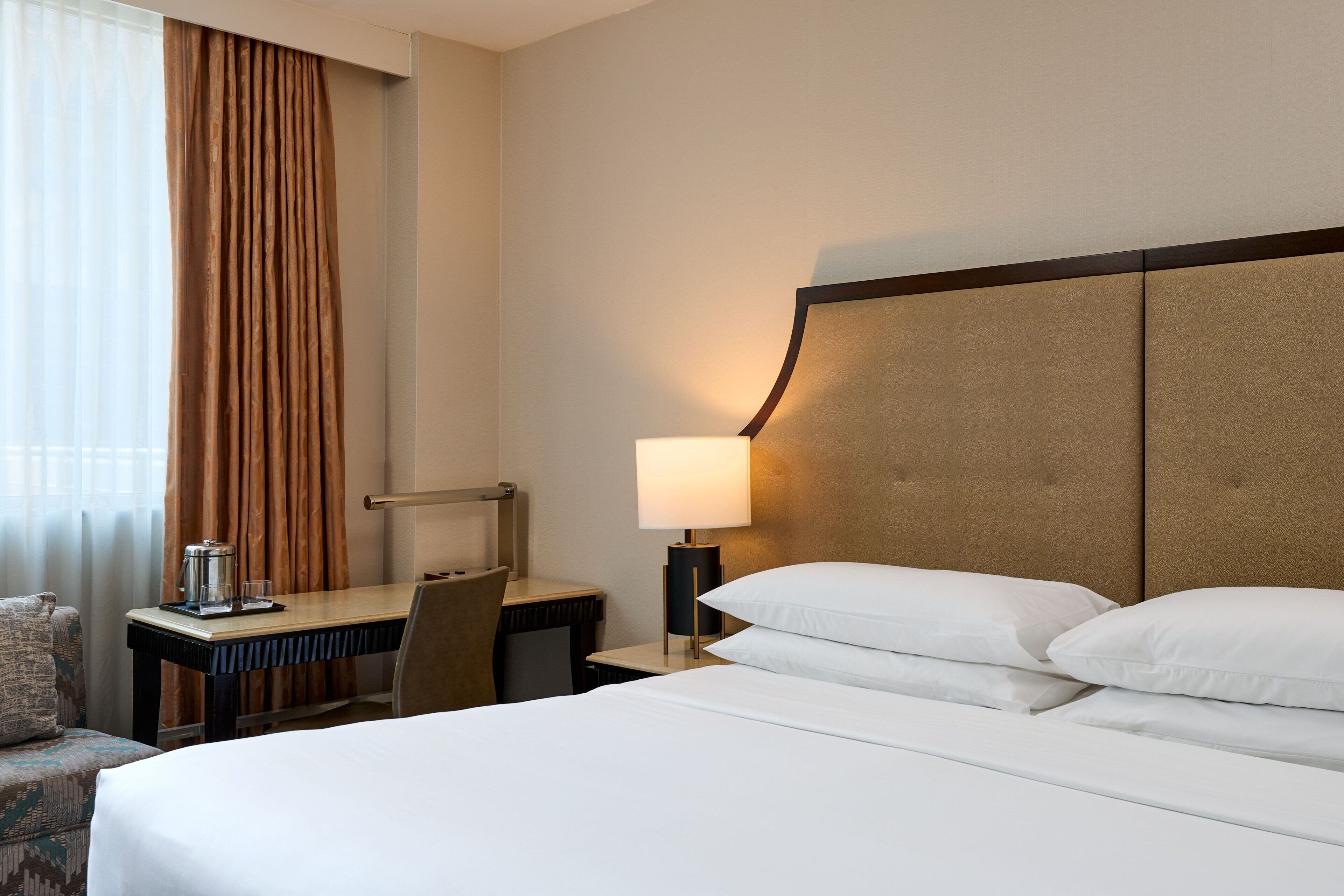 Una habitación de hotel con cama, escritorio, silla, lámpara y ventana con cortinas.