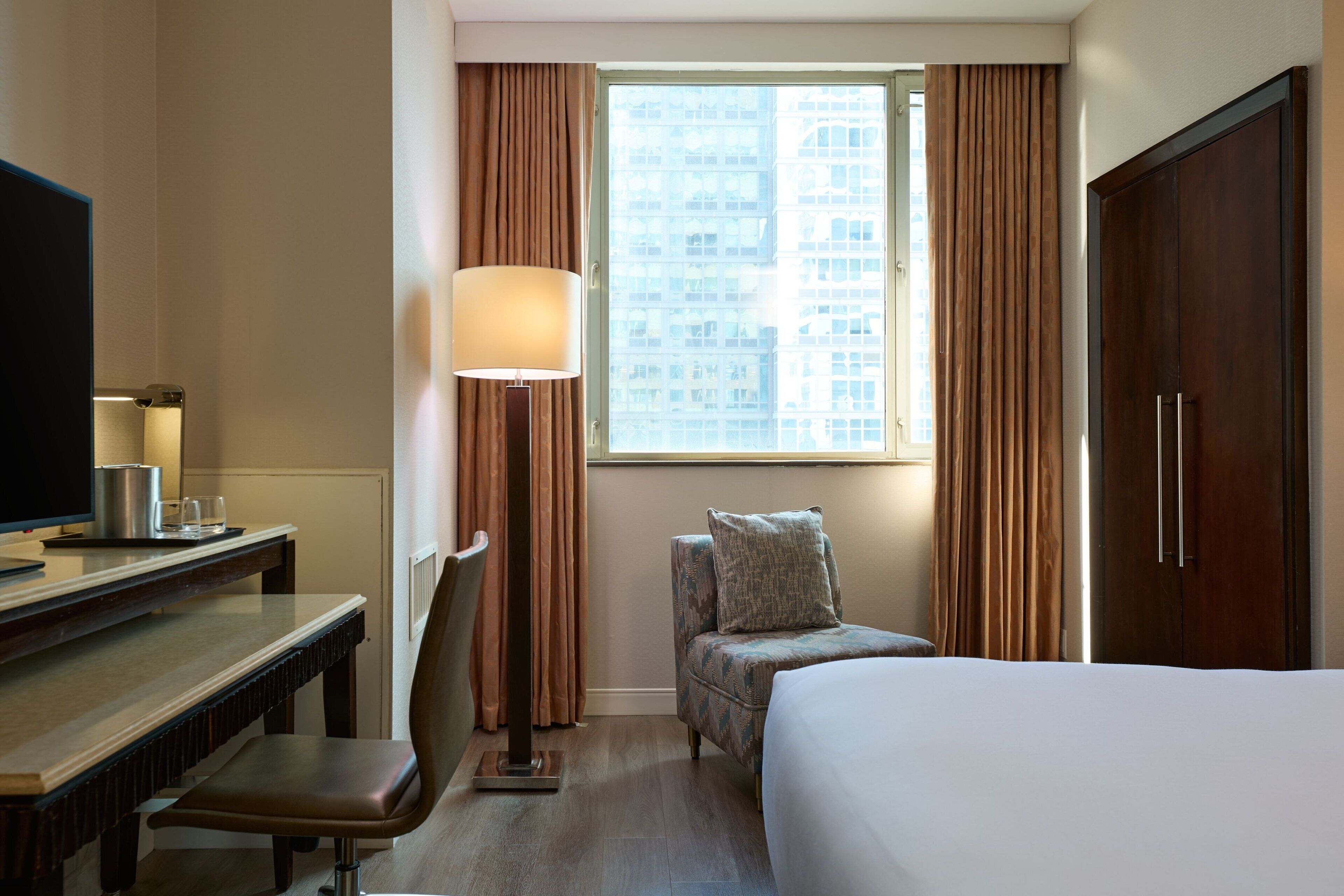A hotel room with a bed, a desk with a lamp, a chair, and a view of a building through the window.