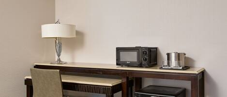 A hotel room with a wooden desk, a chair, a lamp, a microwave, a coffee maker, and a refrigerator.