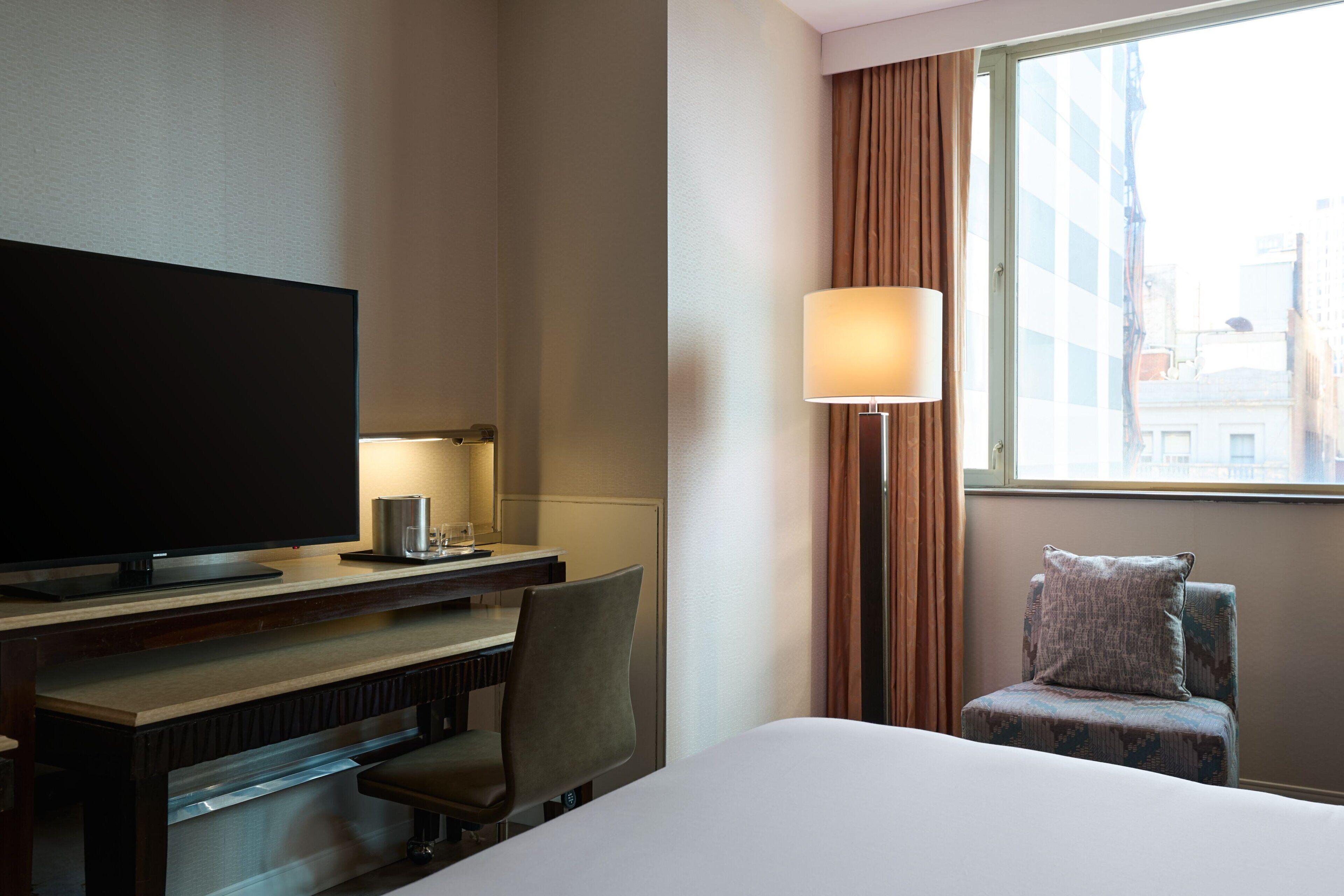 Habitación de hotel con cama, escritorio con televisor, silla, lámpara y ventana con cortinas.