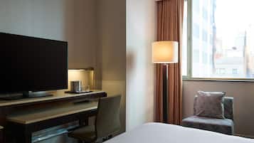 A hotel room with a bed, a desk with a television, a chair, a lamp, and a window with curtains.