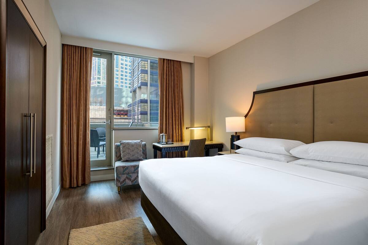 Quarto de hotel com uma cama grande, uma escrivaninha com cadeira, um sofá pequeno e uma janela com cortinas.