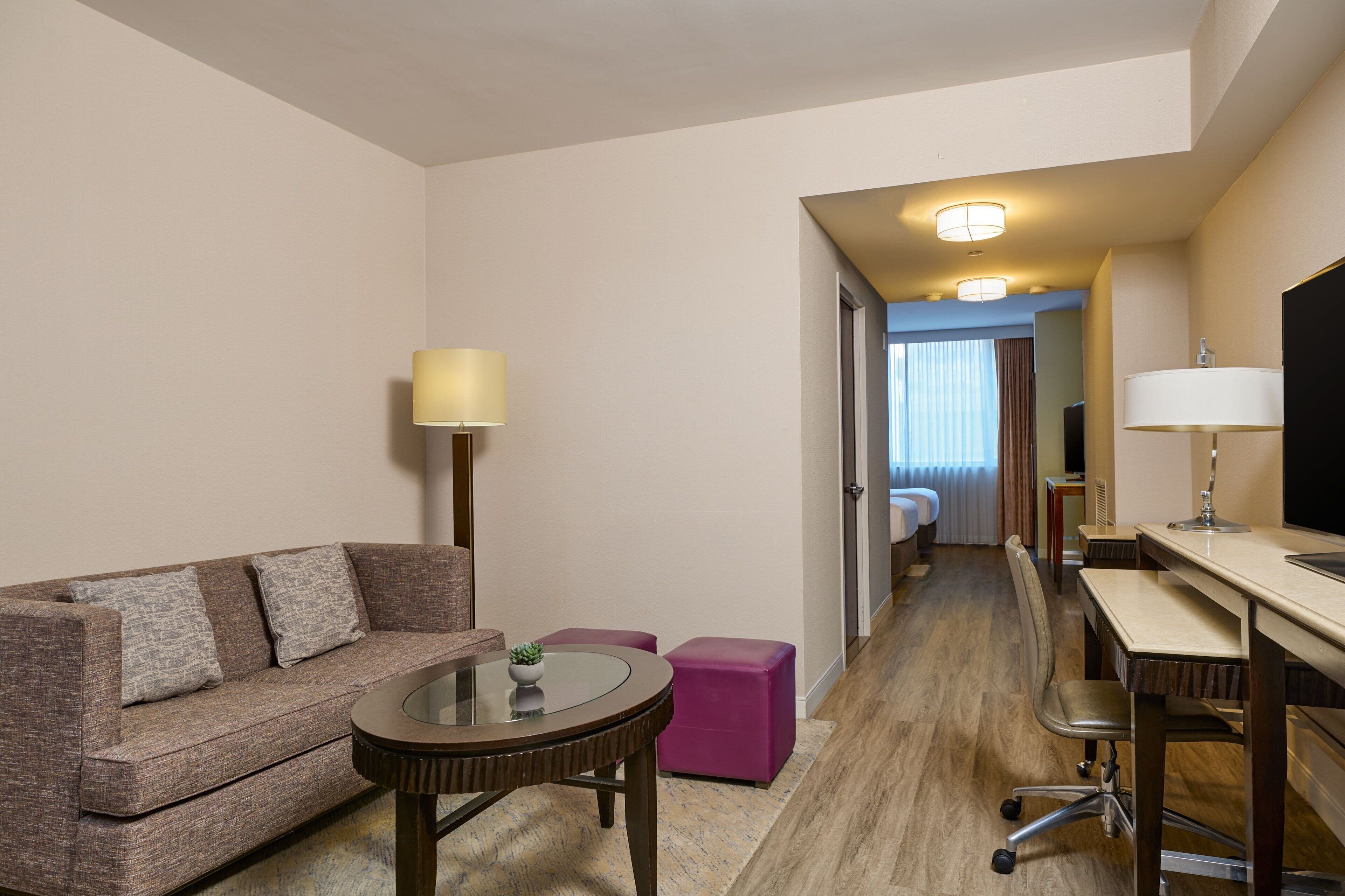 Een hotelkamer met een bank, een klein rond tafeltje, een bureau met stoel en een bed op de achtergrond.