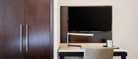 A room with a dark wooden wardrobe, a flat-screen TV mounted on a white desk, and a grey chair.