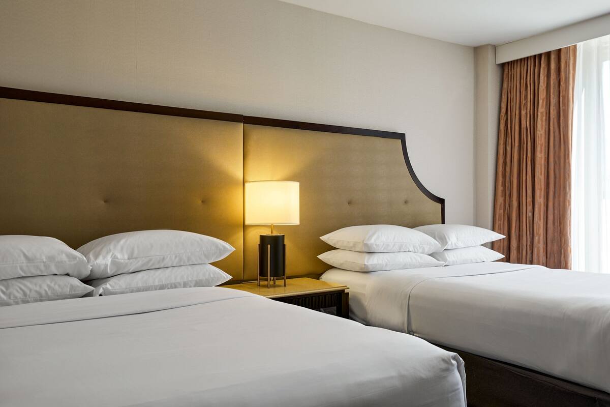 Quarto de hotel com duas camas, uma mesa de cabeceira com abajur e vista para o exterior através de uma janela com cortinas.