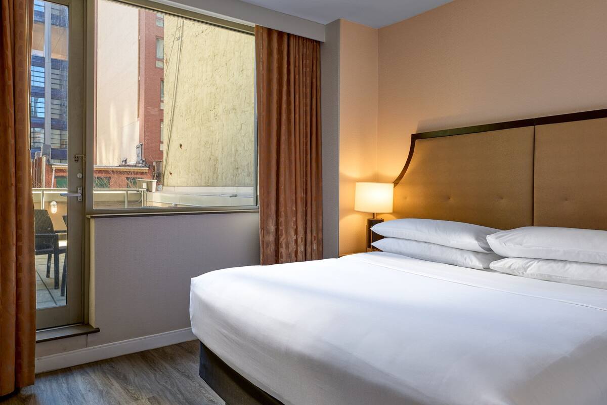 Quarto de hotel com uma cama grande, uma janela com cortinas, mesa de cabeceira com abajur e vista para prédios.