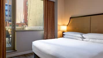 A hotel room with a large bed, a window with curtains, a nightstand with a lamp, and a view of buildings outside.