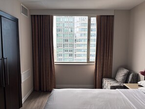 Habitación de hotel con una cama grande, una zona de estar con una silla y cojines, una ventana con cortinas y un armario oscuro.