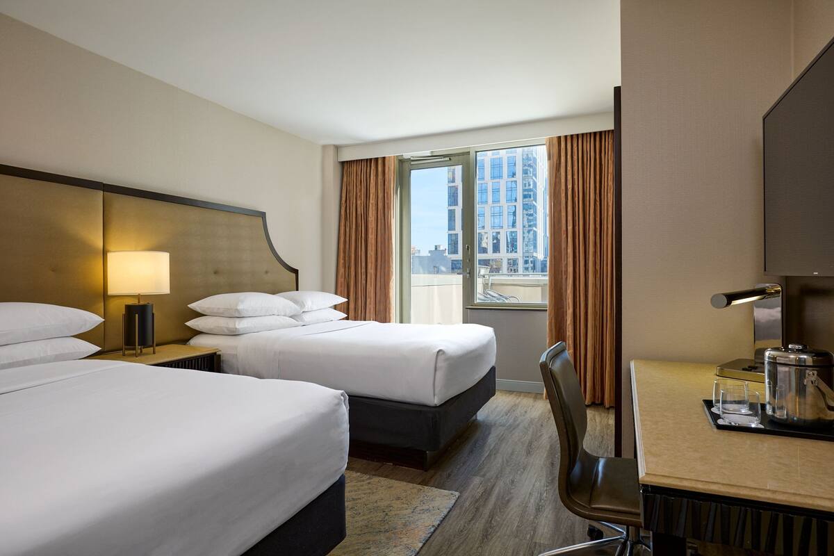 Quarto de hotel com duas camas, uma escrivaninha, uma cadeira, um abajur e uma janela com cortinas.