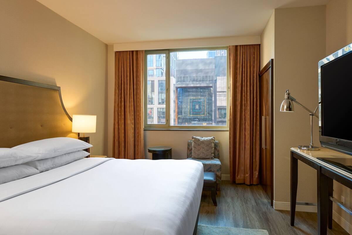 Quarto de hotel com uma cama grande, uma escrivaninha com televisão, uma cadeira, uma janela com cortinas e uma luminária.