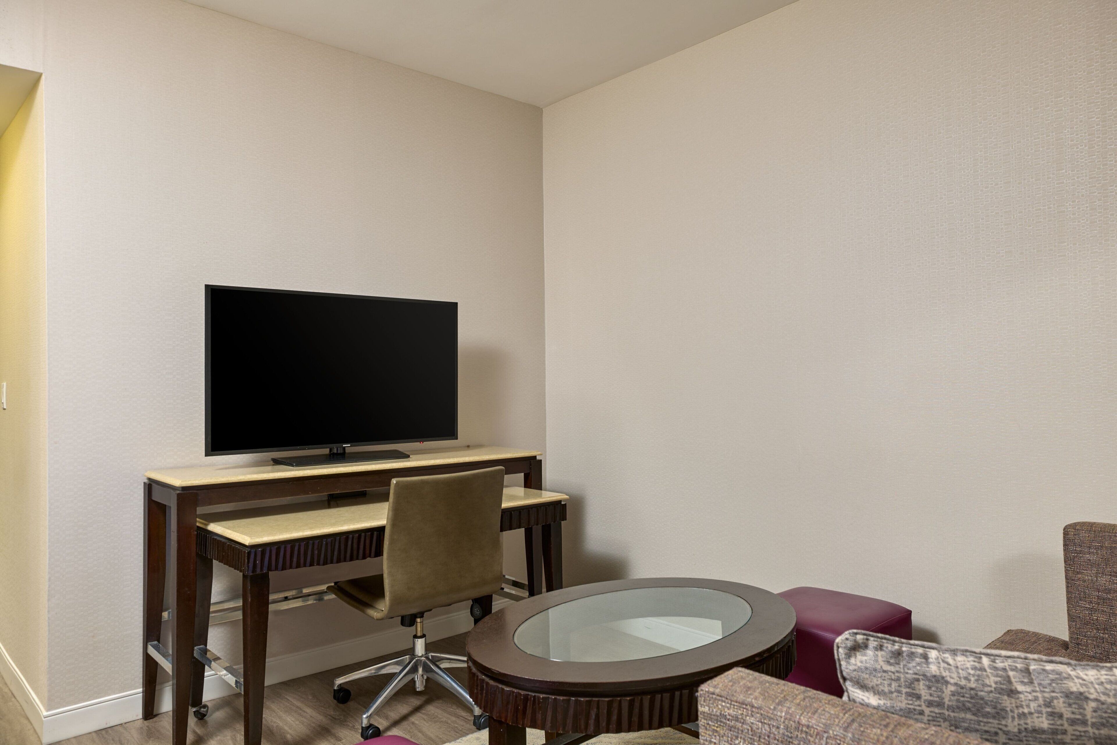 Quarto de hotel com televisão de tela plana fixada em uma escrivaninha de madeira, cadeira giratória de escritório, mesa de centro com tampo de vidro e poltrona estofada.