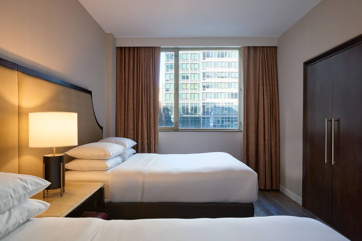 Quarto de hotel com duas camas, uma mesa de cabeceira com abajur, um guarda-roupa e uma janela com cortinas.