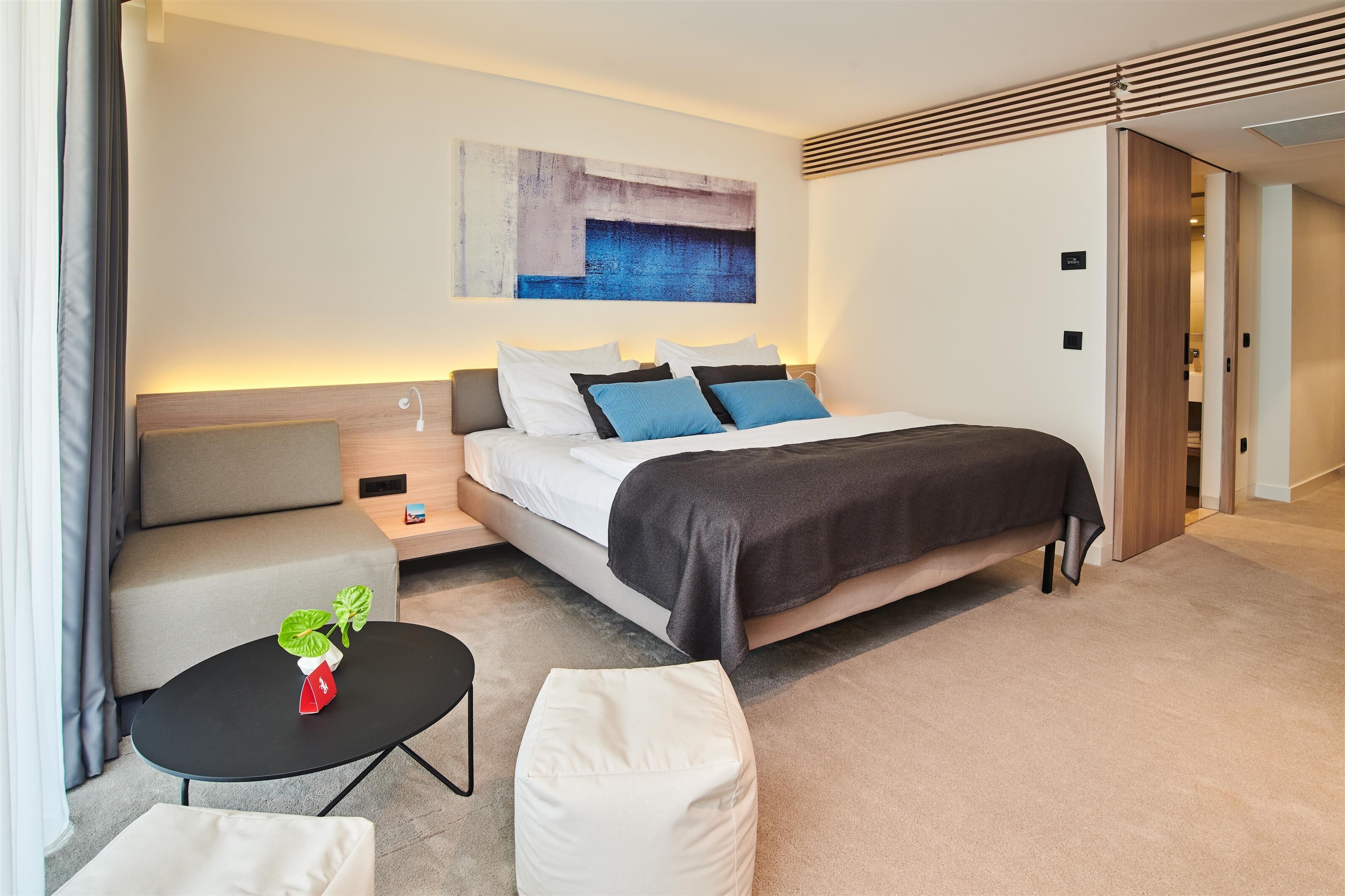 Una camera d'hotel moderna con un letto, comodini, un tavolino con una pianta, un divano e un grande quadro astratto appeso alla parete.